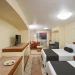 Habitación amplia de hotel con dos camas matrimoniales, área de descanso y comedor, decoración cálida en tonos beige y balcón con vista exterior, ideal para familias o grupos.