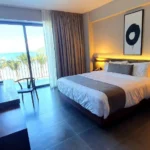Habitación moderna de hotel con cama king size, decoración minimalista y balcón con vista al mar, ideal para disfrutar de una estancia relajante frente a la playa.