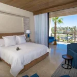 Habitación de lujo con cama king size, balcón con vista al resort y palmeras, decoración moderna en tonos claros y mobiliario elegante para una estancia confortable.