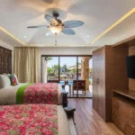 Habitación de hotel con decoración mexicana, dos camas dobles con colchas florales, balcón con vista a la alberca y detalles de madera que brindan un ambiente cálido y acogedor.